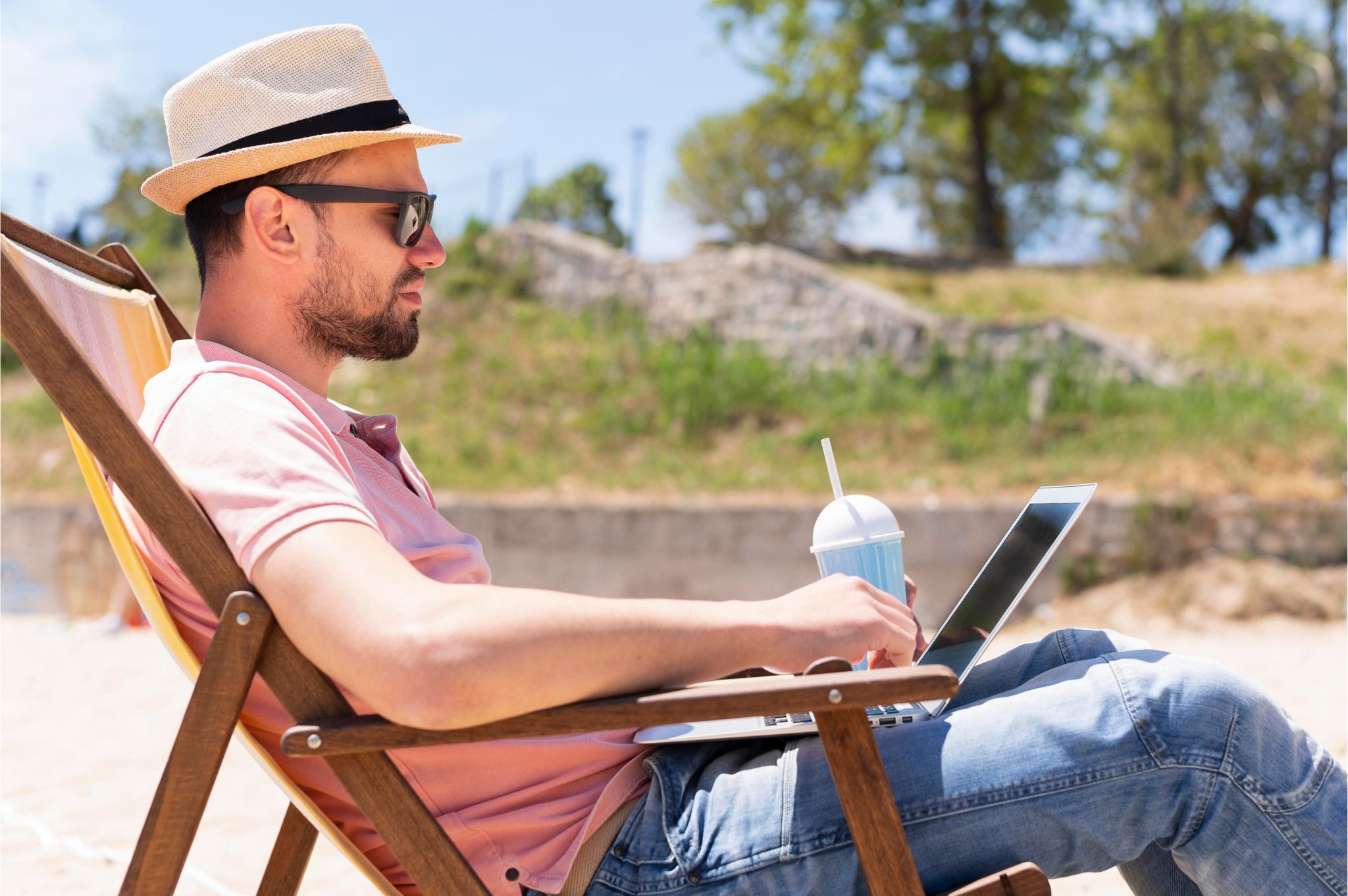 Digital nomad man working on a laptop while relaxing on a beach chair with a drink