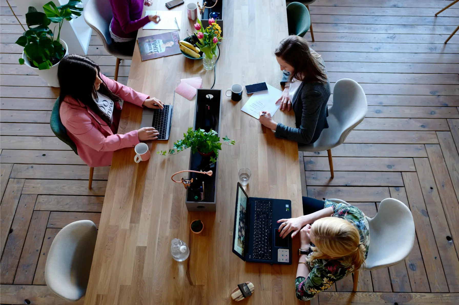 Top-down view of a bright coworking space with four people working at a long wooden table, laptops, notebooks, coffee mugs, and plants.