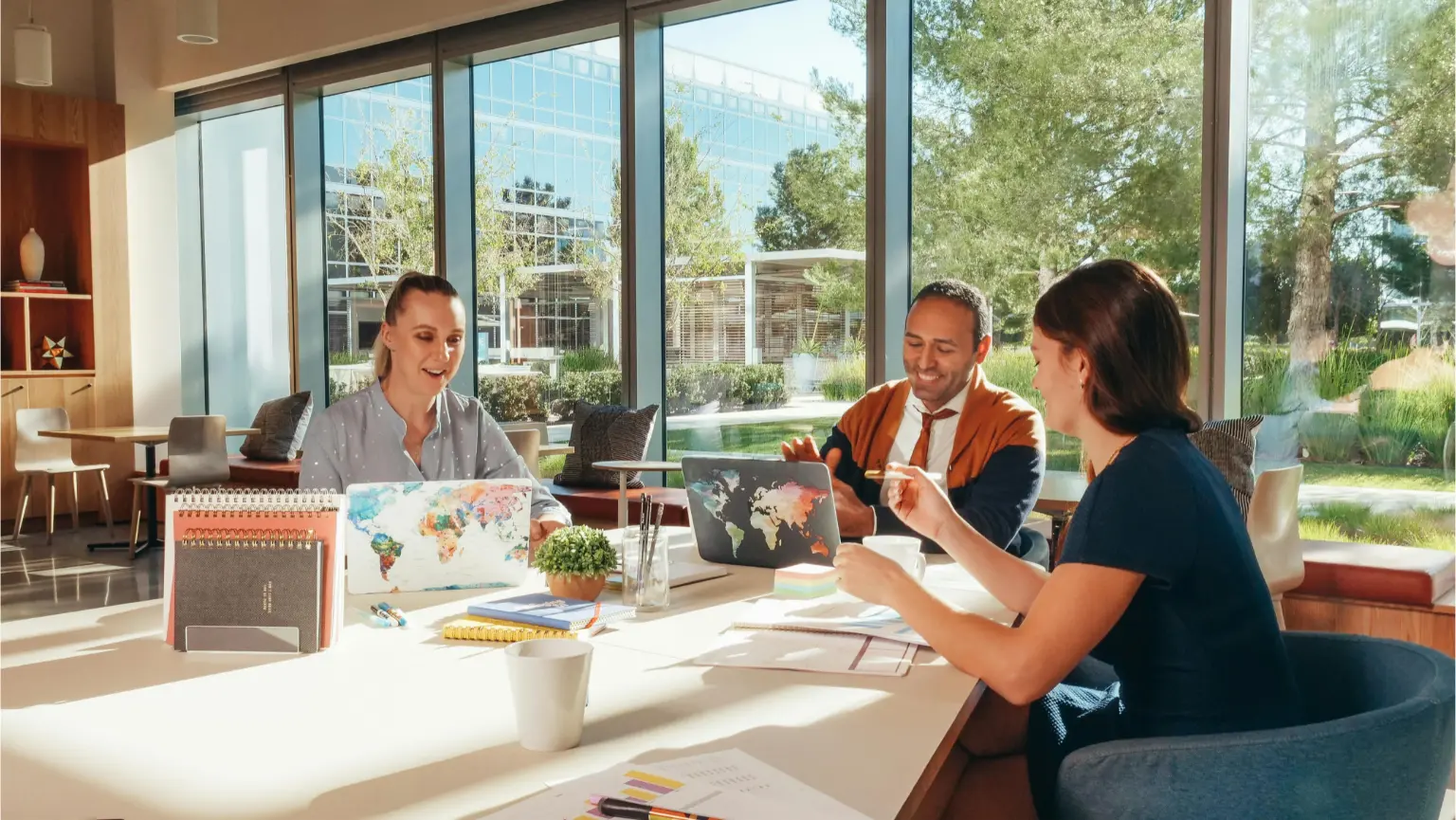 Daytime coworking space with three people collaborating at a large table near floor-to-ceiling windows, laptops, notebooks, and coffee.